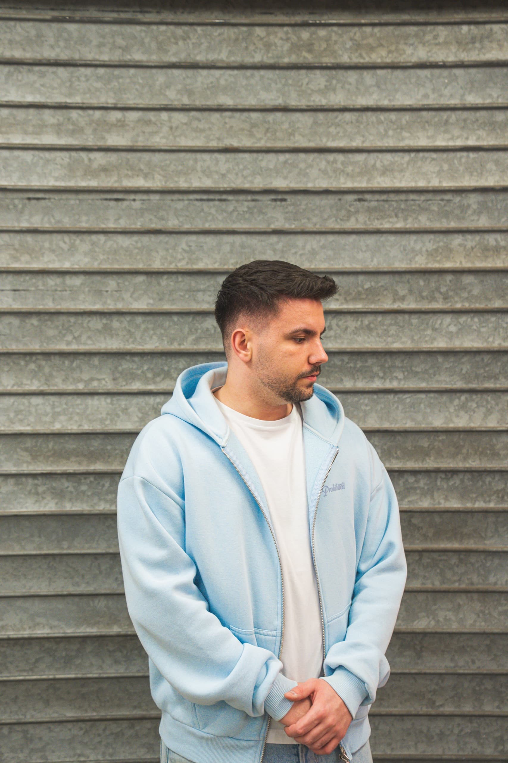 DJ MVM Portrait vor Wellblech in hellblauem Hoodie